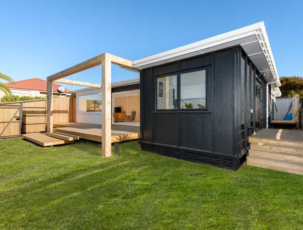 Exterior view of a compact dark-clad single-storey building in a landscaped backyard, with a white low-pitched roof, timber-framed covered deck and outdoor entertaining area, and timber steps and walkway. Large windows reveal a simple interior seating area. A neighbouring suburban house and fencing are visible in the background.