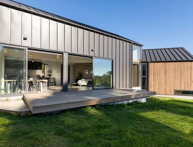 Contemporary single-storey residence with a long, dark metal-clad upper facade, extensive floor-to-ceiling glazing, and a projecting timber deck. A smaller timber-clad wing with a dark pitched metal roof sits to the right. The glazed facade reveals an open-plan kitchen and living area, while the house opens onto a lawned outdoor area.