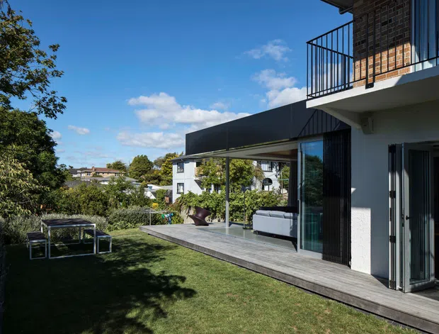 Exterior view of a contemporary multi-level home opening onto a landscaped lawn and timber deck. The facade combines white rendered walls, dark brickwork, extensive glazing, slim black-framed folding doors, and a projecting dark metal-clad roof or canopy. A covered outdoor living area contains seating, while an upper balcony has a simple black metal balustrade. Mature planting and neighbouring homes are visible beyond the garden.