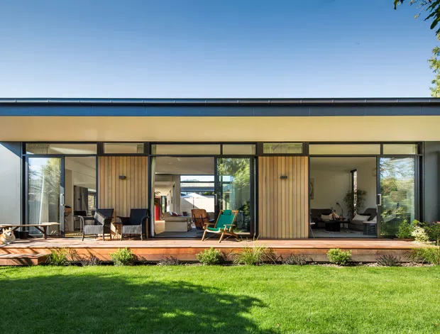 A wide exterior view of a single-storey contemporary home with a long, low-profile roof, extensive floor-to-ceiling glazing, timber-lined wall panels, and a raised timber deck opening onto a lawn. The open plan interior and outdoor furnishings are visible through the glazing, with mature landscaping at the edges.