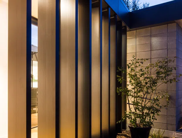 A dusk exterior architectural photograph of a modern courtyard or narrow entry garden. Tall bronze-toned vertical metal fins line a glazed facade, creating a strongly rhythmic screened elevation with warm interior lighting behind it. A dark flat roof connects to a light grey panelled wall, while gravel paving, low ornamental plantings, and a potted tree define the landscaped walkway. The composition emphasizes vertical fins, concealed lighting, and contemporary geometric detailing.