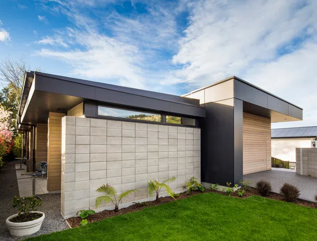 Exterior view of a modern single-storey residence with a strongly articulated flat roof, projecting cantilevered eaves, dark metal cladding, timber-toned wall sections, and light concrete block privacy walls. A landscaped front garden and paved entry path frame the facade.