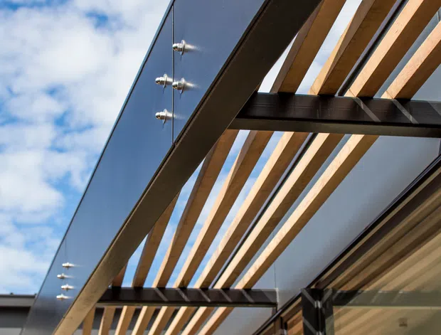 Upward-looking exterior architectural detail showing a contemporary pergola or covered outdoor walkway attached to a building. Repeated warm timber slats project from dark steel beams beside a dark grey metal-clad fascia, with large glazed doors or windows below. Strong sunlight creates linear shadows across the facade, and blue sky is visible above.