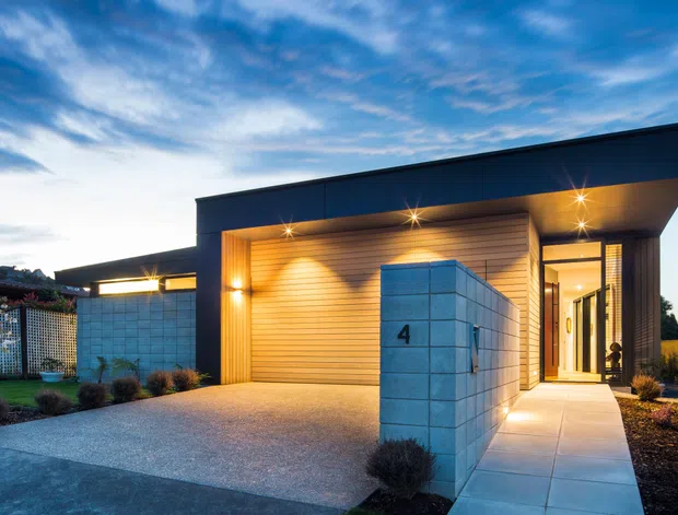 Dusk exterior view of a contemporary single-storey residence with a low-slung flat roof, dark metal-clad facade, warm timber-look horizontal wall lining, and a prominent concrete block entry wall marked with the number 4. The illuminated garage and glazed entry sit beside a broad textured driveway and landscaped planting beds.