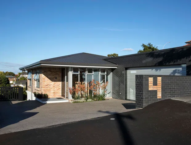 Single-storey residential home viewed from the driveway, with a low-pitched dark metal roof, warm brick facade, white-framed glazing, and a dark brick-clad attached garage. The driveway and garage dominate the foreground, with a small covered entry and landscaped planting near the front windows.