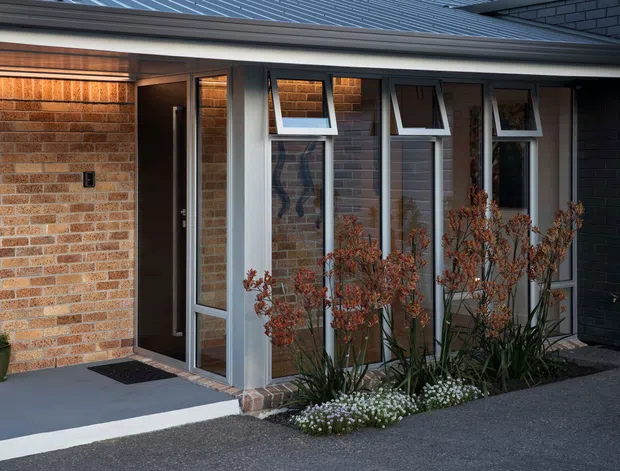 Exterior view of a contemporary single-storey entry facade. The composition features warm exposed brick walls, a dark metal pitched roof with broad eaves, a dark front door, and a glazed entry corridor with multiple narrow awning windows in light grey frames. Low planted garden beds sit beside a concrete path and entrance platform.