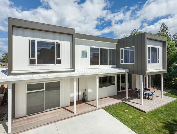 A contemporary two-storey residential house viewed from the rear, featuring a crisp white and charcoal-grey facade, projecting upper-level volumes, vertical corrugated cladding, large windows and sliding glass doors. The lower level opens onto timber decking and a covered patio with outdoor dining furniture, while a concrete path and lawn occupy the foreground. The home has a flat or very low-pitched roof form and a bright suburban setting.