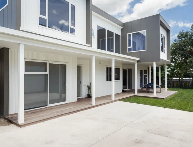 Exterior view of a contemporary two-storey residential building with crisp geometric volumes, white vertical cladding, dark grey upper-level panels, large windows, and a covered timber-decked veranda supported by slim white posts. A broad concrete driveway or forecourt occupies the foreground, with a small lawn and outdoor seating to the side.