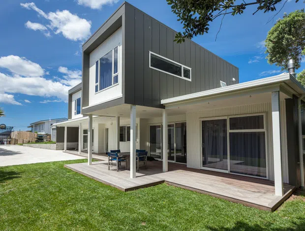 Exterior view of a contemporary multi-level residential house with a contrasting dark vertical-panel upper volume, white rendered and weatherboard-like lower sections, extensive glazing, covered outdoor living areas, and a timber deck opening onto a lawn. The home has a strong geometric form with projecting upper-storey elements and a low-pitched roofline.
