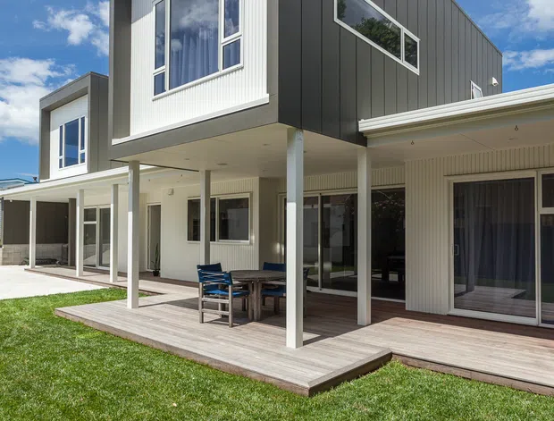 Exterior view of a contemporary two-storey house with a sharply angular flat-roofed form. The upper level is clad in dark vertical panels with contrasting light fibre-cement or ribbed cladding around the windows. Large glazed openings and sliding doors connect the ground floor to a broad timber deck and covered outdoor entertaining area. A manicured lawn occupies the foreground under a bright blue sky.