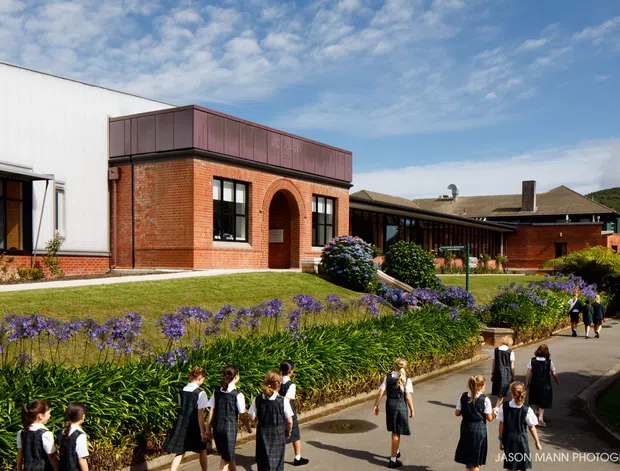 Exterior view of a red-brick school or institutional campus with a contemporary addition, arched brick entry, dark metal roof elements, landscaped garden beds, and students walking along paved paths under a partly cloudy sky.