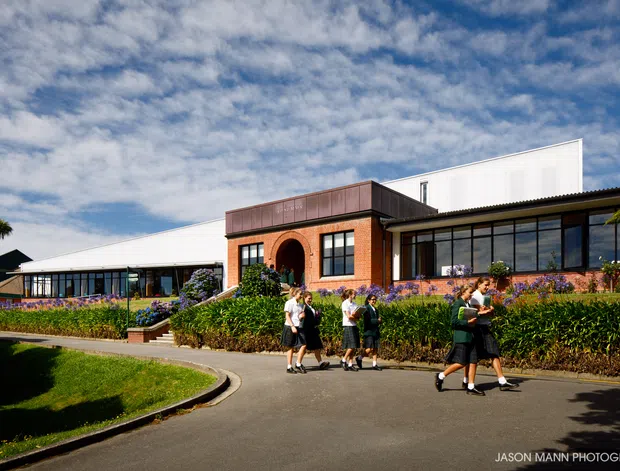 Exterior view of a low-rise institutional or commercial building with red brick walls, large glazed sections, white corrugated metal wall panels, and a dark flat-roofed entrance volume. A paved driveway and landscaped garden border the building, with people walking outside.