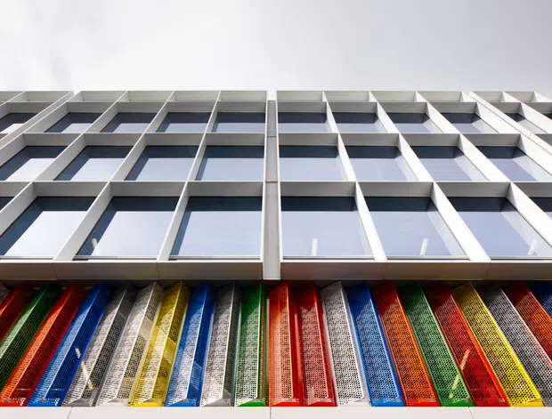 Upward-looking architectural photograph of a contemporary multi-storey facade. Repetitive white-framed window bays form a strong geometric grid above a vivid lower facade of multicoloured perforated or patterned panels, creating a bold contrast between the neutral upper structure and saturated lower detailing.