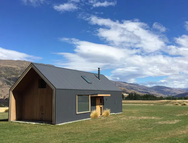 A compact modern cabin set in an open rural valley, with a steep gabled charcoal metal roof, dark corrugated metal cladding, and a warm timber-clad gable end and entry surround. The simple single-storey building has a roof skylight, chimney, and large front window, with dry grass and rugged hills forming the surrounding landscape beneath a bright blue sky.