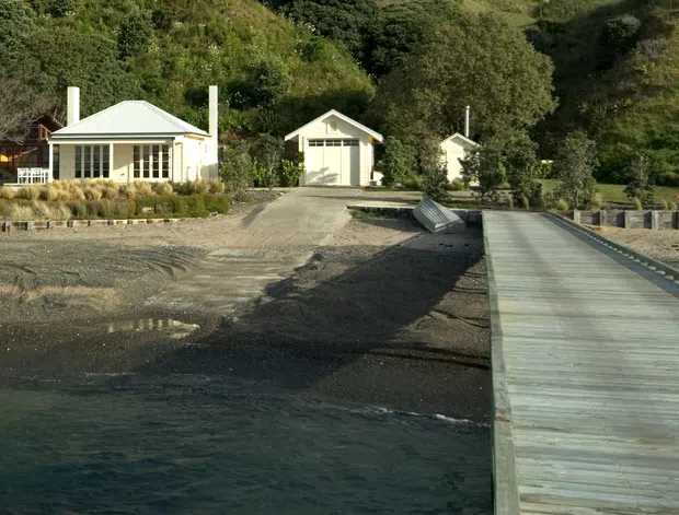 A small waterfront residential compound viewed from the water, with light-coloured single-storey buildings set against a steep, densely vegetated hillside. The main cottage has a low hipped roof, verandah-like glazing and pale vertical cladding, with two smaller outbuildings behind it. A long timber jetty extends from the shore into the foreground water, while gravel landscaping and rock edging define the shoreline.