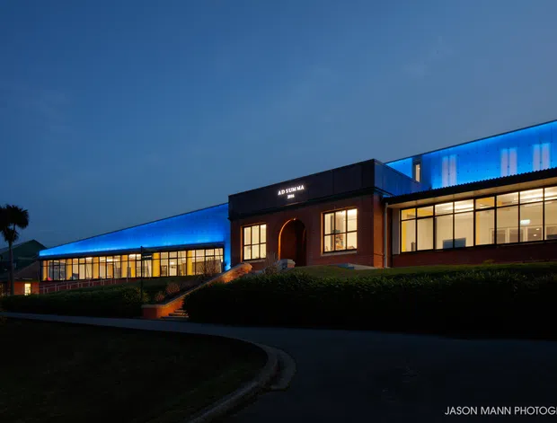 A dusk exterior photograph of a low-rise modern commercial or institutional building. The composition features long glazed wings, a central dark brick entrance pavilion with an arched doorway, illuminated interiors, and vivid blue linear facade lighting along the roofline. A landscaped hedge, steps, curved driveway, and palm silhouettes occupy the foreground.