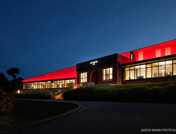 Night-time exterior view of a long, low modern building on a landscaped site. The facade combines extensive warm-lit glazing, dark masonry or cladding, and striking red illuminated wall panels beneath a flat roofline. A central entrance with steps and a curved driveway are visible.