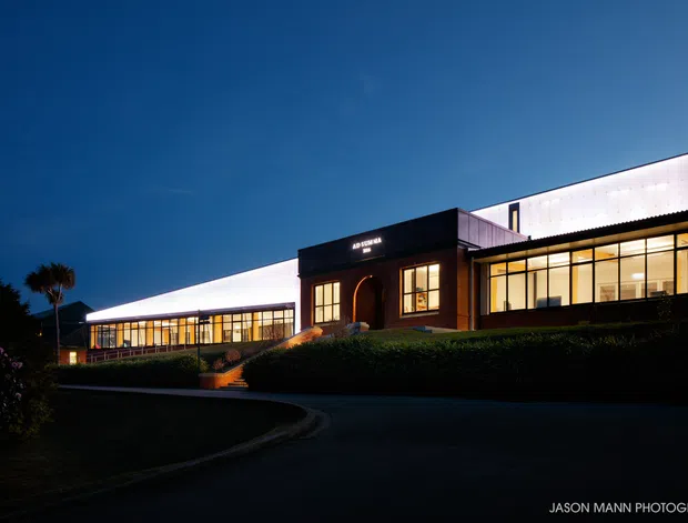Night-time exterior photograph of a low, modern institutional or commercial building composed of interconnected rectangular volumes. The dark central entry block has a recessed arched doorway and illuminated signage, flanked by extensive floor-to-ceiling glazing. Bright translucent upper wall or roof elements create a strong horizontal glow, with landscaped hedges, a curved driveway, steps, and silhouetted palms in the foreground.