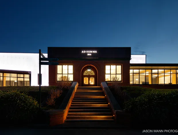 Night-time exterior view of a modern commercial building with a dark brick central entrance, illuminated stairway, arched doorway, large glowing windows, and contrasting white and dark horizontal wings. The sign above the entrance reads “AD SUMMA 2016.” Landscaping frames the approach.