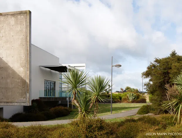 Exterior view of a modern concrete and white-clad building set within landscaped grounds. The composition highlights a tall exposed concrete feature wall, projecting roof forms, glazed openings, sculptural yucca-like plantings, a pedestrian path, and contemporary street lighting beneath a partly cloudy sky.