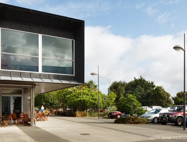 Exterior view of a contemporary, box-shaped commercial or mixed-use building with extensive glazed upper-level windows, dark metal cladding, and a projecting canopy. The ground level opens onto a paved pedestrian forecourt with outdoor café-style tables and chairs, landscaped planting, modern street lamps, and a parking area with cars. Bright daytime setting with a clean urban-suburban character.