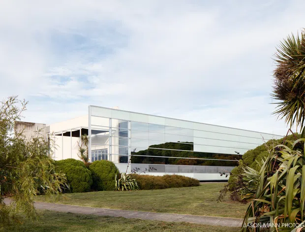 Exterior view of a low, modern building set within a landscaped garden. The facade features extensive reflective horizontal glazing, slender metal framing, and crisp white structural volumes, with sculptural plants and manicured greenery in the foreground.