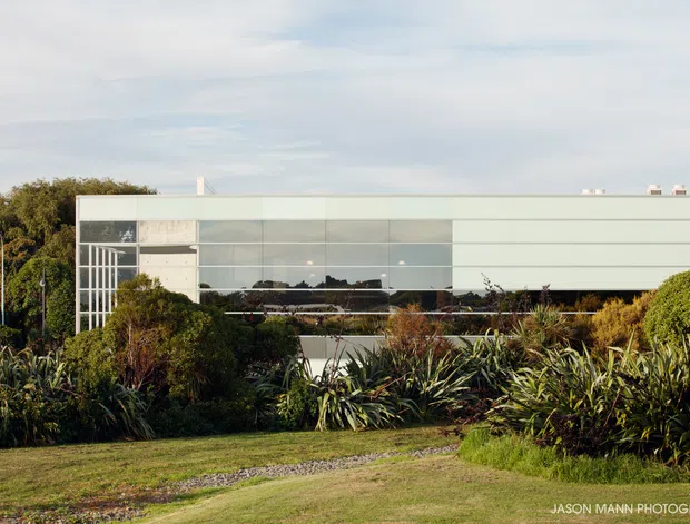 Exterior view of a low-rise modern building set within a lush landscaped garden. The long, rectilinear facade combines broad horizontal pale cladding panels with extensive reflective glazing and a glazed corner or entrance zone. Dense coastal-style planting and a lawn occupy the foreground, while the flat roofline and minimal detailing create a clean contemporary appearance.