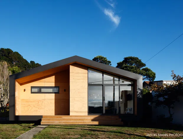 Exterior view of a compact contemporary single-storey house with a dramatic sloping roof, warm timber-clad facade, extensive floor-to-ceiling glazing, and a small timber entry deck. The home is set against a vegetated hillside under a clear blue sky.