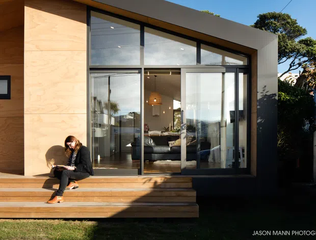 Exterior view of a contemporary residential extension with a warm timber-clad wall, broad black-framed glazing, sliding glass doors, and a timber deck with steps. A living room is visible through the glazing, while landscaped planting surrounds the home.