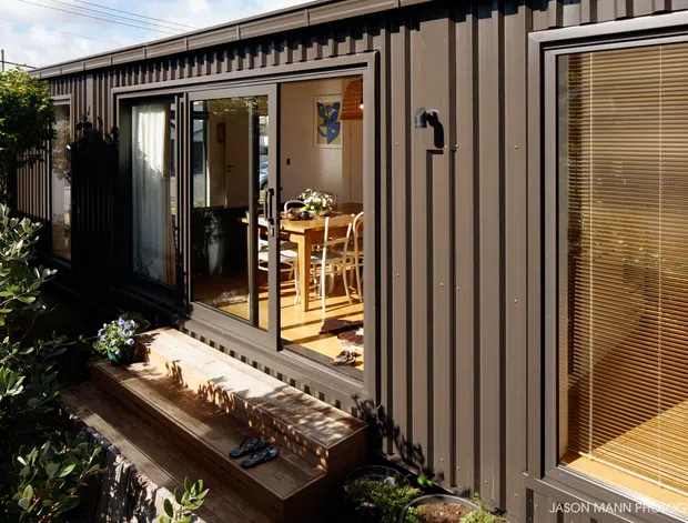 Exterior view of a compact single-storey dwelling clad in dark brown vertically ribbed metal panels. Large glazed sliding doors open onto a timber deck, revealing a bright dining area inside, while a window with timber venetian blinds is visible on the right. Dense planting borders the deck.