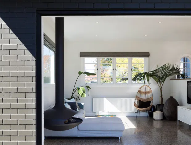 A bright contemporary living room viewed from an exterior paved area through a large open glazed opening. The room features white walls and ceiling, polished terrazzo flooring, a pale sectional sofa, a suspended black sculptural fireplace, indoor plants, a cane lounge chair, recessed downlights, and abundant natural light. The exterior facade is dark painted brick with black metal framing and a low roof overhang.