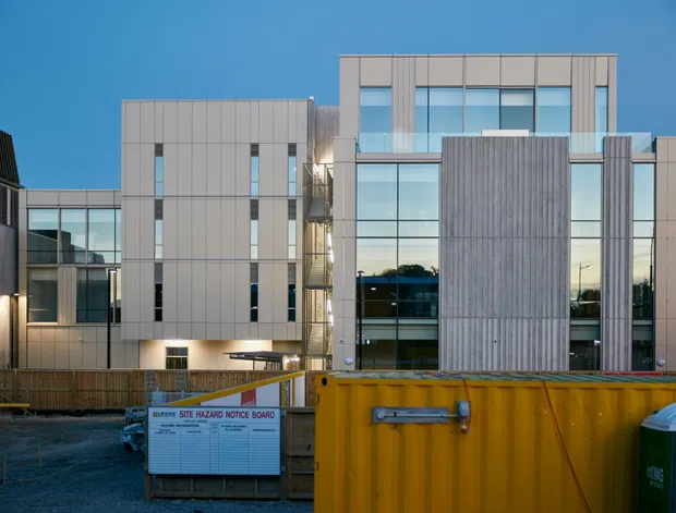 A contemporary multi-storey commercial or institutional building viewed from the street during construction. The facade combines pale vertical cladding panels, extensive floor-to-ceiling glazing, and prominent vertically textured concrete or cement-panel sections. Temporary construction fencing, a site notice board, a yellow skip bin, and portable bins occupy the foreground. The building has a rectilinear flat-roof form with a recessed glazed central section and strong vertical facade articulation.