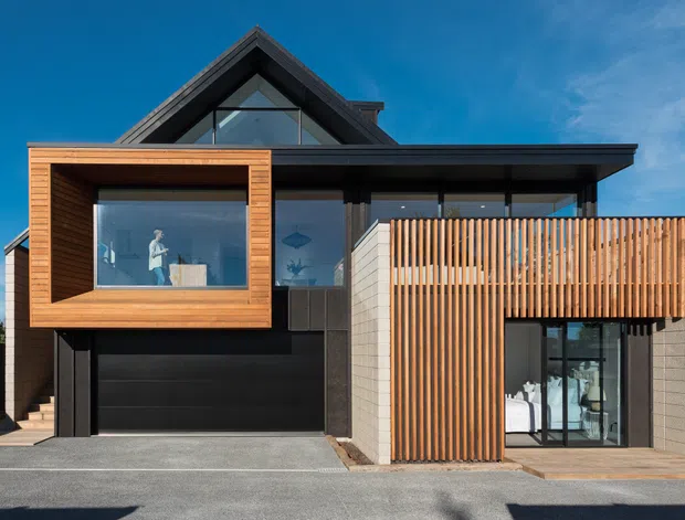 Street-facing exterior view of a contemporary multi-level residence with a steep gabled roof, dark cladding, extensive glazing, a projecting timber-framed upper-level window, and a vertical timber privacy screen over the ground-floor facade. A double garage is integrated beneath the projecting volume.