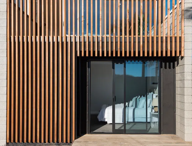 A contemporary exterior facade with vertical timber slat screening over a dark-framed glazed bedroom opening. The slatted screen continues upward to form a privacy balcony or upper-level enclosure, while pale masonry/concrete block walls frame the composition. A timber deck sits at the base, and a bed is visible through the sliding glass doors.