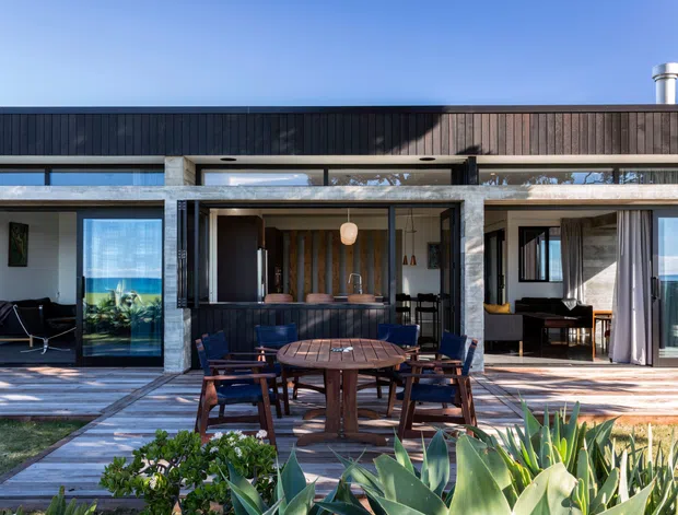 Exterior view of a low, contemporary coastal home with dark vertical cladding, extensive glazing, and a covered outdoor dining terrace. The terrace includes a timber dining table and chairs, while indoor living spaces and a compact kitchen or bar area are visible through the open glazing. A timber deck, paved path, planted garden, and ocean horizon contribute to the relaxed setting.