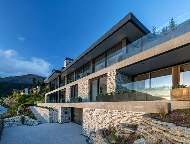 Exterior view of a contemporary multi-level residence built into a sloping mountain site. The building features strong horizontal cantilevered volumes, extensive floor-to-ceiling glazing, glass balustrades, dark metal roof and trim, and extensive natural stone masonry walls. Landscaped terraces and a driveway lead toward the house, with forested mountains visible in the background.