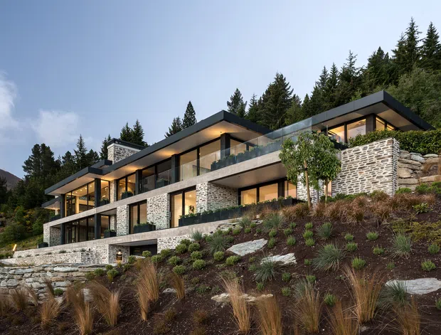 A contemporary multi-level hillside residence set into a landscaped, forested mountain site. The house features broad floor-to-ceiling glazing, strong horizontal cantilevered roof planes, extensive natural stone masonry walls, dark metal window frames, and glass balustrades. Terraced planting and stone retaining walls integrate the building into the sloping terrain.