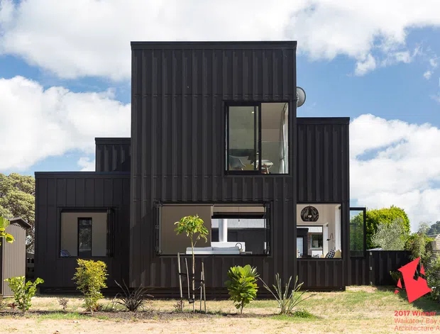 Exterior view of a compact contemporary multi-level home clad in dark vertically ribbed panels. The asymmetrical facade has projecting and recessed box forms, large rectangular windows, and a glazed ground-floor opening. A simple landscaped front garden sits beneath a bright partly cloudy sky.