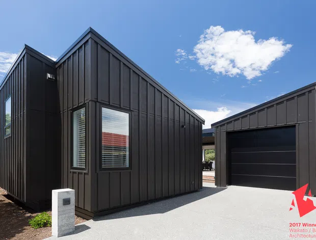 Exterior view of a contemporary dark-clad residential building with a steep gabled main volume and matching detached garage. Vertical black cladding, narrow windows with blinds, a paved driveway, and landscaped surroundings are visible under a bright blue sky. The image includes a small architectural award graphic in the lower-right corner.