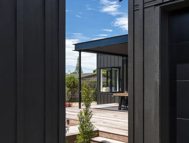 Exterior view through a narrow passage between two dark-clad building volumes, leading to a timber deck and covered outdoor area. The structures feature crisp modern lines, black vertical cladding, projecting roof edges, and dark-framed glazing and doors. Small landscaped planting and gravel appear beside the deck under a bright blue sky.