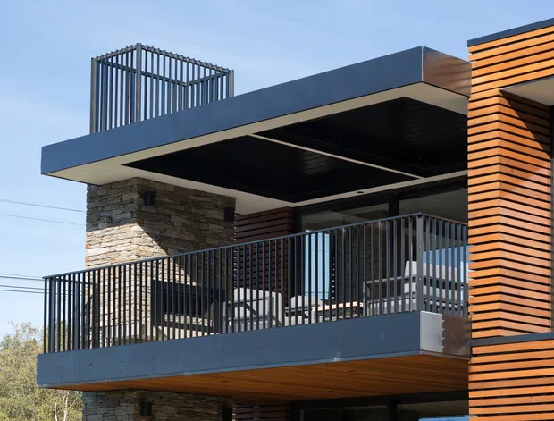 Contemporary multi-level residential exterior with projecting balconies, dark metal railings, a flat roof or cantilevered canopy, warm horizontal timber cladding, and a substantial stacked-stone feature wall. The image focuses on the building facade and outdoor balcony spaces against a clear sky.