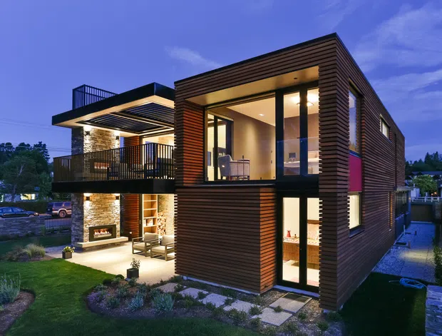 Twilight exterior view of a contemporary multi-storey home with a strongly rectilinear form, extensive glazing, projecting balcony, flat roof planes, and horizontal dark timber-style cladding. Warm interior lighting reveals an open living area and upper-level room, while landscaped gardens and paved paths surround the house.