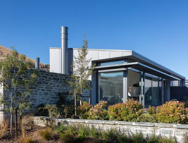 Exterior view of a low-profile contemporary house set into a dry, sloping landscape. The building combines dark stone masonry walls with pale metal-clad volumes, extensive floor-to-ceiling glazing, projecting flat roof planes, and prominent metal chimney flues. Layered stone retaining walls and landscaped planting define the foreground.