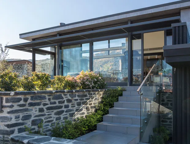Exterior view of a contemporary multi-level house set into a landscaped, sloping site. The facade combines extensive floor-to-ceiling glazing, dark vertical cladding, exposed concrete roof edges, deep horizontal sun-shading elements, and a broad exterior stair with glass balustrades. Low stone retaining walls and raised planting beds frame the entrance terrace. Mountainous terrain is visible through the glazing and in the background.