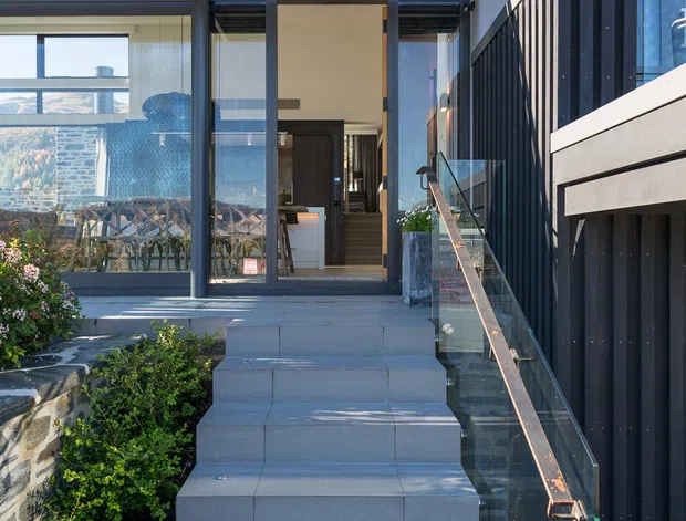 Exterior view of a contemporary multi-level residence approached by broad grey tiled steps. The entry is framed by expansive glazing, a dark metal slatted pergola, dark vertical cladding, and a clear glass balustrade with timber-toned handrail. Landscaping and a stone retaining wall border the stairs, while the open doorway reveals a small portion of the interior.