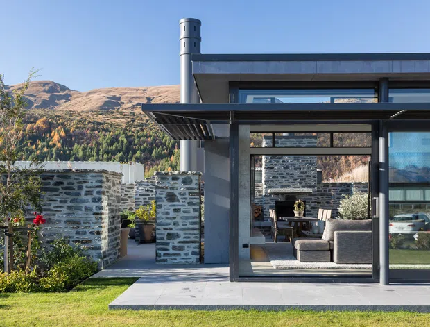 Exterior view of a contemporary single-storey residence with dark metal framing, large glazed openings, a projecting flat canopy, and a cylindrical metal chimney. A landscaped courtyard with low dry-stone walls and outdoor planting sits in front of the home, while the interior living area is visible through the glazing. Mountainous terrain forms the distant backdrop.