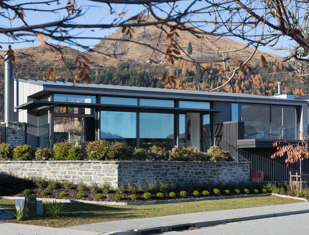A low, contemporary single-storey residence set against a dramatic mountainous landscape. The home features a predominantly dark grey and charcoal exterior, extensive black-framed glazing, projecting flat roof planes, and a sheltered entry or outdoor living area. A low rough-stone retaining wall, landscaped garden beds, and a paved verge define the street frontage.