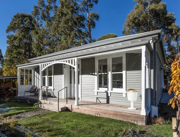 Single-storey cottage-style house viewed from the front garden, featuring grey horizontal weatherboard cladding, white-framed windows and doors, a low pitched roof with broad eaves, and a covered front veranda with decorative timber brackets and posts. Mature trees and landscaped lawn surround the home.