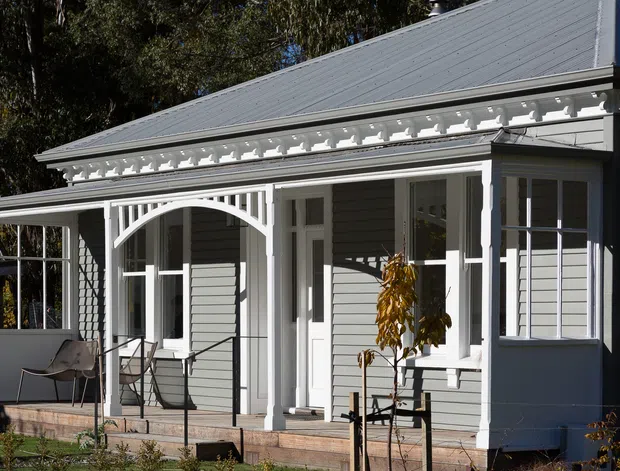 Exterior view of a single-storey cottage-style house with a grey weatherboard facade, white timber-framed windows and doors, a covered front veranda, ornate decorative eaves trim, and an arched veranda detail. The pitched metal roof and traditional detailing are clearly visible, with minimal landscaping in front.