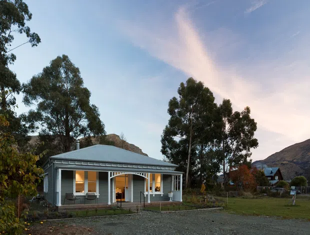 Exterior view of a single-storey cottage-style home in a spacious rural or semi-rural setting. The house has a symmetrical front verandah, pale horizontal weatherboard cladding, white trim, a low hipped corrugated metal roof, and tall multi-pane windows and doors. Mountains and mature trees surround the property, with gravel ground and open lawn in the foreground.